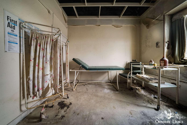 An abandoned and decaying first aid room featuring a treatment bed and a rolling medical cart. A "First Aid" poster is visible on the left wall