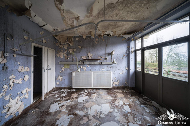 An abandoned hospital room shows extensive decay with peeling purple paint and debris covering the floor and walls. The ceiling is crumbling,