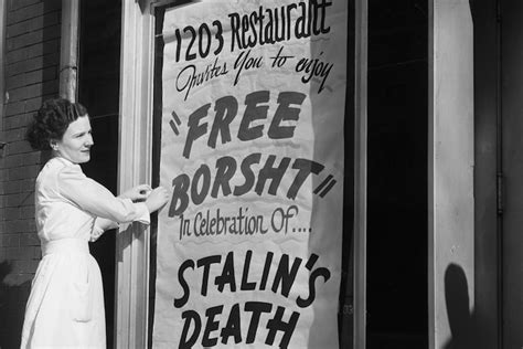 A black and white photo of a woman posting a large banner "free borsht in celebration of Stalin's death" on her restaurant window