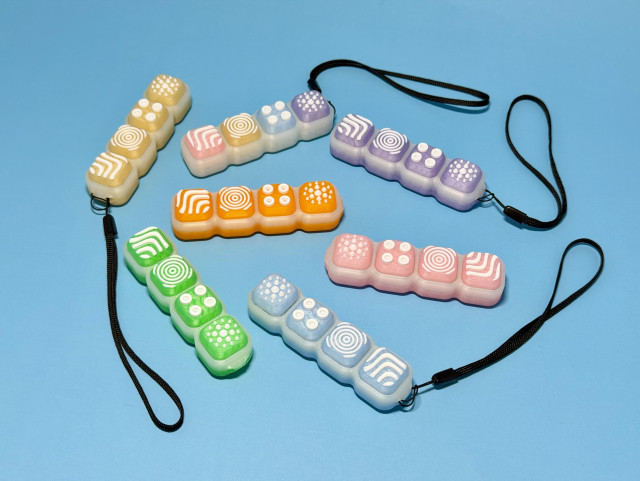 In a baby blue background: keyboard clicker fidgets with wrist straps. Designed and printed to be a throwback to the early 2000’s translucent designs from that era. Available in rainbow, yellow, green, orange, purple, pink and blue. 

The tops of each of the keycaps is raised and textured for sensory input. 