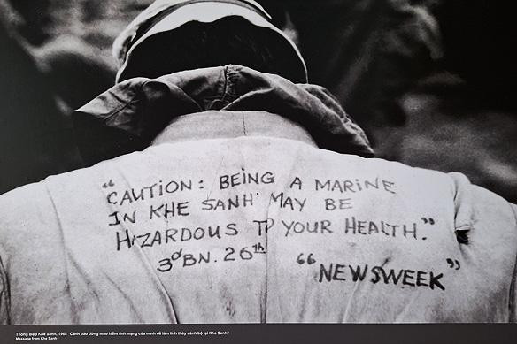 ‘Message from Khe Sanh.’ [Khe Sanh Combat Base Museum]
Black and white image of a person's back, wearing an army jacket with handwritten text. The text reads: "CAUTION: BEING A MARINE IN KHE SANH MAY BE HAZARDOUS TO YOUR HEALTH." and "3rd BN. 26th" with the word "NEWSWEEK" in quotes below. The man is seen from the back, wearing a hat, with the jacket's collar up.