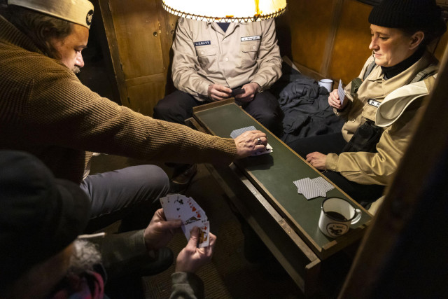 Eine Gruppe von vier Spielern an einem Tisch. Enge, dämmriges Licht. Sie spielen Karten. Eine Kaffetasse steht auf dem Spieltisch, die Spieler tragen eine Art Seemanns-Uniform.