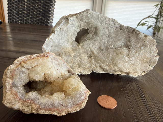 Two geodes on a table, with a penny for scale. The back one is big and heavy, with one penny-sized hole that contain clear crystals. 
The front one is smaller, and yellow. The hole is larger, and crystals seem bigger. 