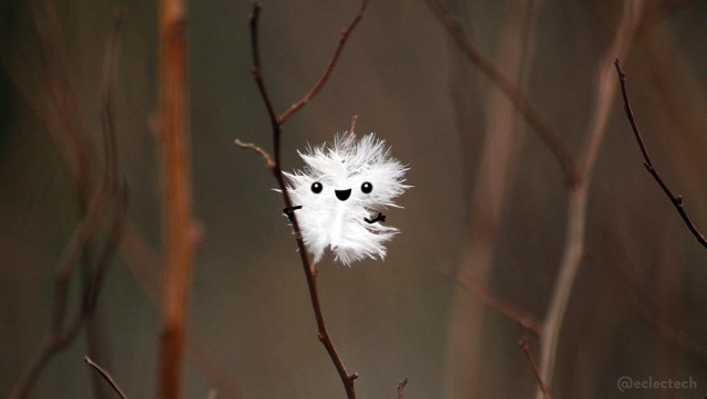 A photo of a small white fluffy feather stick on a thin branch against a brown (and sticky) blurred backdrop. The feather has a happy face drawn on, and little arms, one of which holds on to the branch, the other is waving at you.