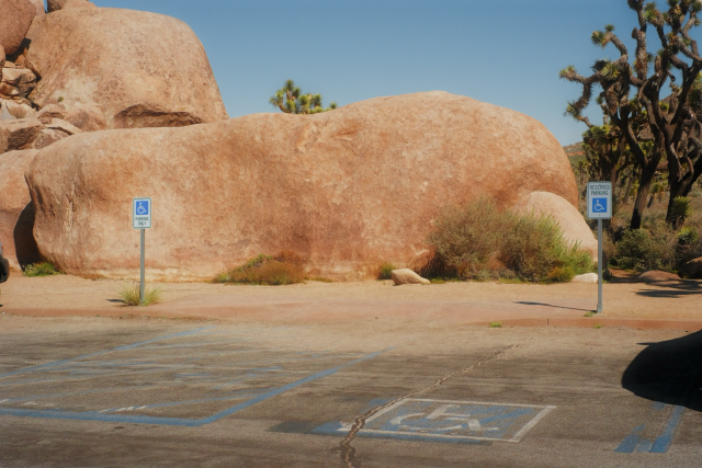 A photo of a parking lot at Joshua tree state park. There are two blue reserved wheelchair parking signs in front of the large rocks.the dichotomy of the signs against the rocks is vaguely humorous. 