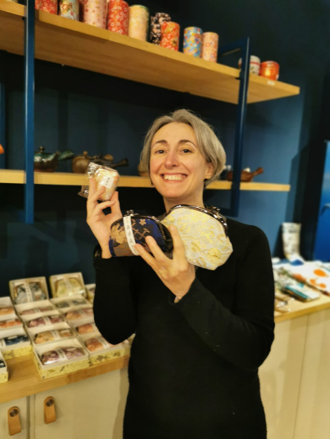Elisabetta, a person with short gray hair, stands in a tea shop with a cheerful expression. She is wearing a black top and holding two items: a small, elegant tea tin with a floral design and a transparent bag filled with loose tea. Behind her, wooden shelves display an assortment of colorful tea tins, boxes, and accessories, contributing to the shop's cozy and organized ambiance.