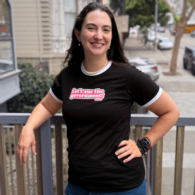 Person wearing EFF's new black and white ringer tee. Features the text "Let's Sue The Government!" in white and pink.