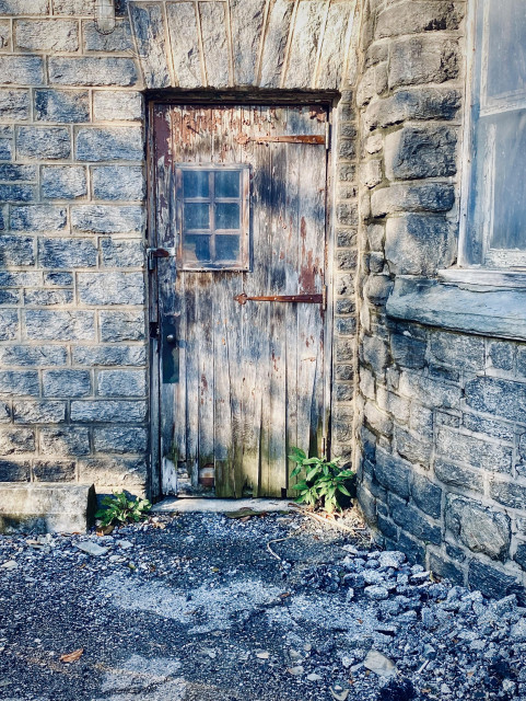 Very old wood door with rusted antique strap hinges and small viewing panel of 6 small square panes is installed above the handle. It’s very crude, weathered and warped. The entrance is at the corner inside a long straight wall of the building to left, unseen, and a curving wall to the right. Building is made of mortared blocks of stone. An old asphalt sidewalk or parking pavement is almost crumbled up leading to the door. It looks sinister but maybe used to be magnificent before decades of decay. 