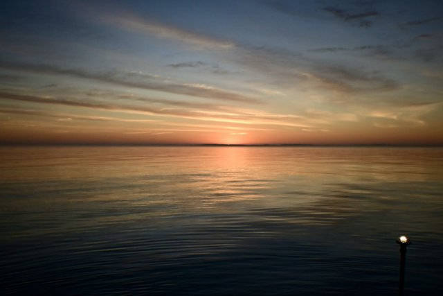 Reflections of orange on the water with the sun rising on the horizon. Clouds scattered and opening to a blue sky. 