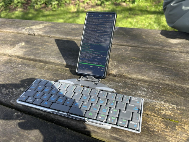 Photo of a vertical Pixel set atop a Stowaway Bluetooth keyboard, on a wooden picnic table. The screen is showing a tmux session in termux, running the BSSG build process.