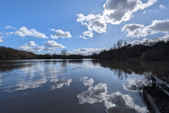 A reasonably still calm lake reflects the blue cumulus cloud dotted sky above. The sun is just poking through a cloud and is clearly visible as a spot of bright on the lake.