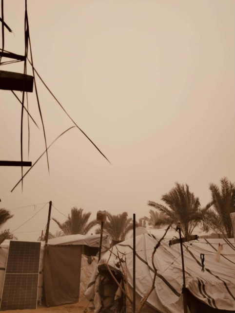 An outdoor camp scene under a hazy, sand-filled sky, featuring multiple tents, palm trees, and a solar panel, with visible wires and supports.