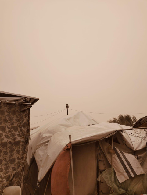 Dust-filled sky over makeshift structures of white tarps and patterned fabric, suggesting a refugee camp or similar temporary settlement.