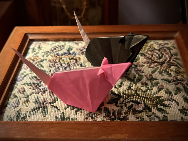 Two small 3D origami rats on the cushioned jewelry box lid, one pink and the other one black, both with a long white tail. They’re lit from the left, the tail side, by the lamp. The room is dimly lit.