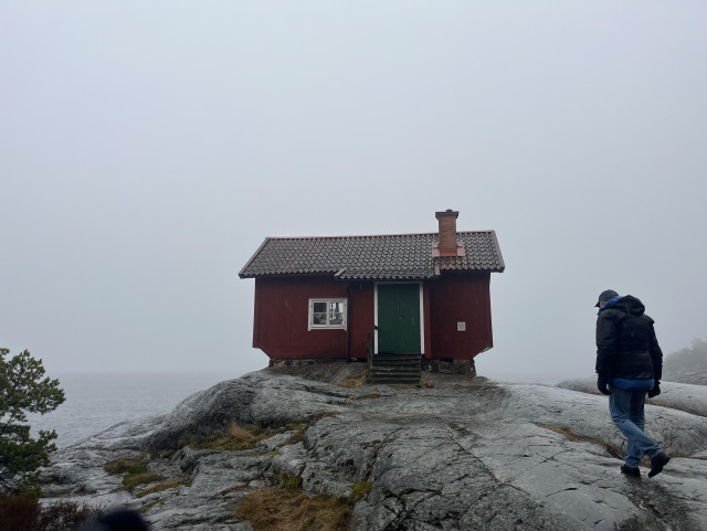 Dimma, grå himmel och ett dis över havet. En person går över blöta klippor upp mot ett litet rött hus (Albert Engströms ateljé) som har vita knutar och grön dörr och ligger precis vid havet.