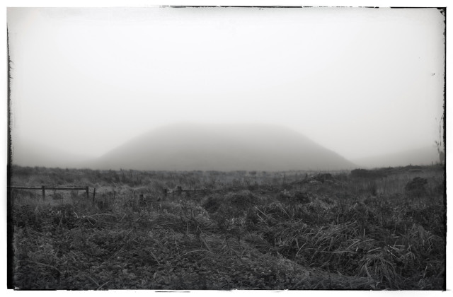 A monochrome photo of a field of grassy chaparral and a large hill in the distance, shrouded in fog. There's a long fence cutting through the field but nothing else. It is bleak and dismal.