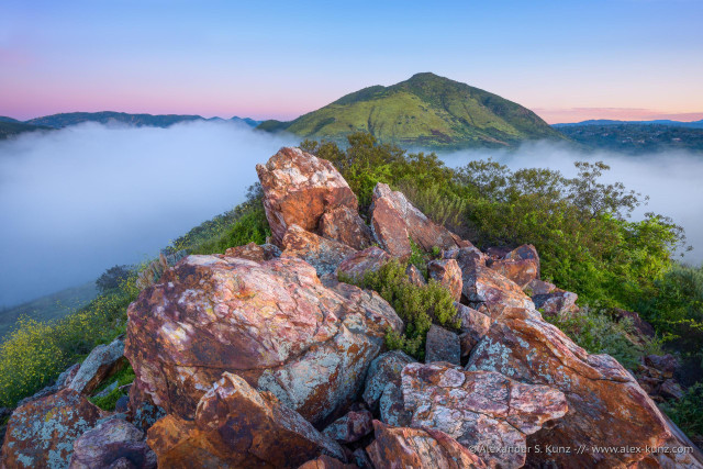 Color landscape photo with a prominent pile of irregular shaped red brown rocks in the immediate foreground. The rocks are on a ridge above a valley that is filled with morning fog. Further back is a rounded, solitary hill or small mountain with green slopes. The sky is clear and the scene is illuminated by the indirect light of the pre-dawn sky.
