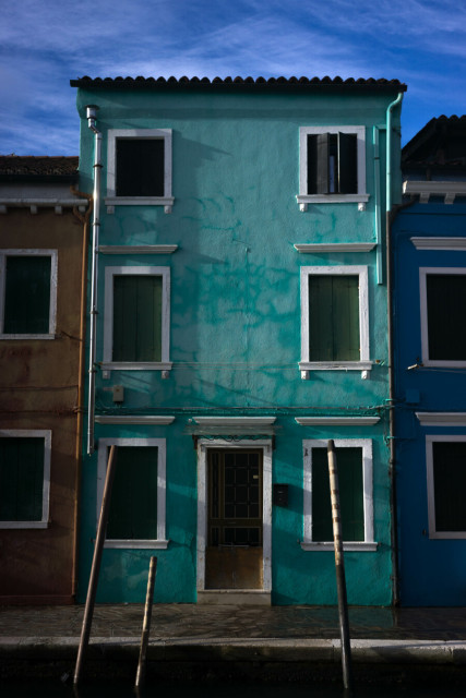 An aquamarine house, six windows, white frames, dark shutters, stands foursquare between shorter yellow and blue neighbours under a blue sky. Three thin mooring poles standing in an unseen canal seem almost to prop it up.