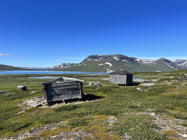 Two old wooden huts on tundra landscape by a lake. First one is very rotten, a ruin, the other one (on stelts) seems to be still in use, there’s a bar with a lock at the door. Mountains with patches of snow in the background. Blue water, bluest sky. 