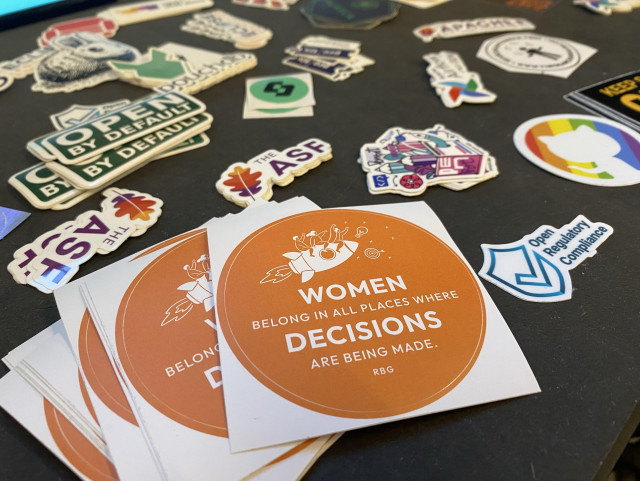 A collection of various colorful stickers scattered on a black surface, featuring a prominent round sticker with the text "WOMEN BELONG IN ALL PLACES WHERE DECISIONS ARE BEING MADE. RBG."