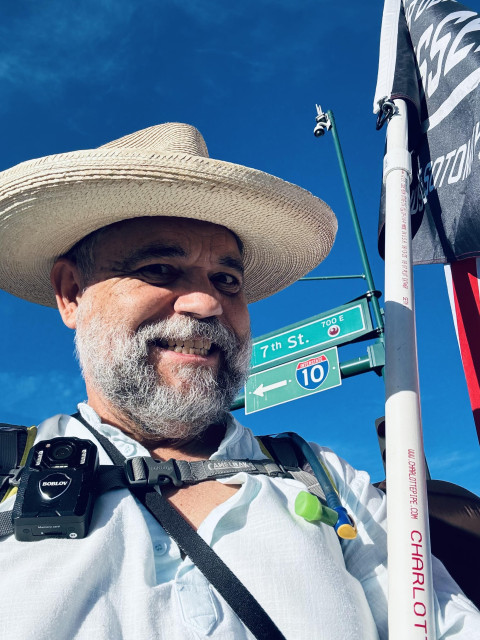 Selfie, wearing a wide-brimmed hat and a body camera. Blue sky and a portion of my protest flag in the background. 