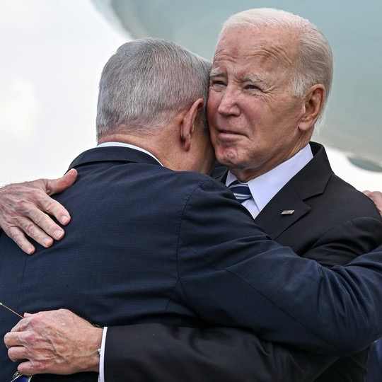 U.S. President Joe Biden embraces Israeli Prime Minister Benjamin Netanyahu during an official meeting, both men wearing dark suits. Biden’s face is visible over Netanyahu’s shoulder, showing a solemn expression as they hold each other closely.