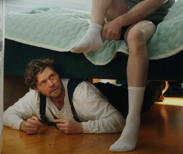 Picture of the video showing the Enshittificator under the bed of a (male?) person putting his socks on, while the Enshittificator is about to attack and cut a hole in the sock.