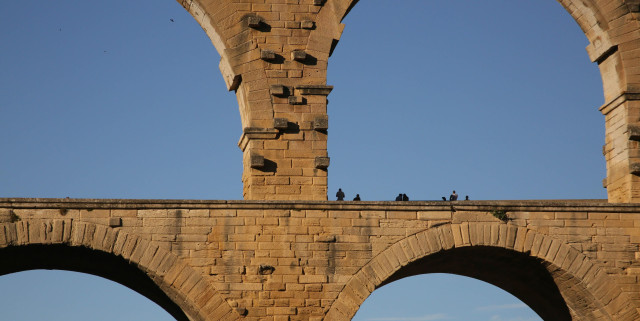 Partial view of a Roman aqueduct made of yellow stone, with blue sky behind and small figures standing on it.