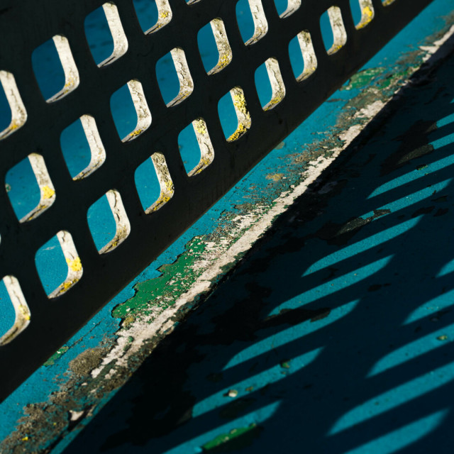 Angled photo of a grid of rounded squares in a slice of darkness and their corresponding elongated blue holes in shadow. This is an outdoor table tennis table, the white holes in the rigid net have cracks and lichen around their edges and the blue table surface beneath is peeling, revealing green and white paint beneath.