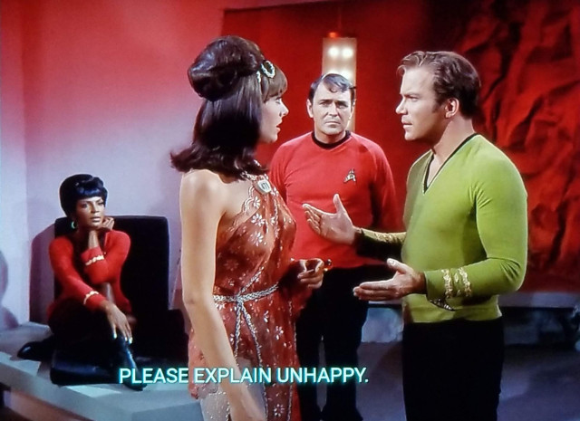 Star Trek The Original Series scene. We're in a room on an "away mish" and the room is like half wall and half cave, which Star Trek loves to do. The cave half is lit in red. Kirk is standing and talking to a woman in an up-do haircut wearing a shimmery dress and showing a lot of arm and shoulders. Scotty is standing behind them looking suspicious. Uhura sitting in the background listening intently and catching tea. Kirk's hands are extended out in a 'what gives?' motion. Closed caption reads, "Please explain unhappy."