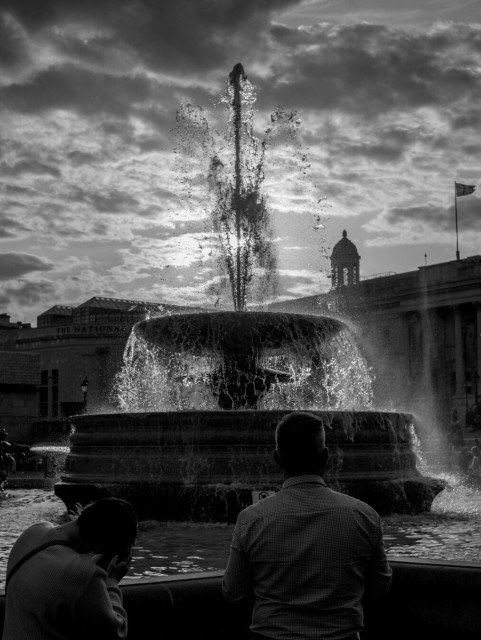 Monochrome photo of a large round fountain backlit by a bright, but cloudy, sky, meaning that the water seems to glow. In front of the fountain, looking towards it, are a couple of figures, near-silhouettes, outlined in light.