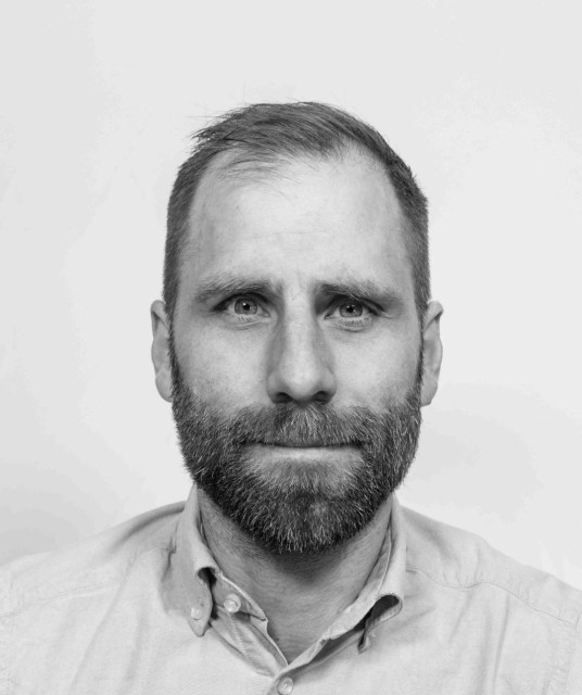 A black and white close-up self-portrait of a middle-aged man with short hair, a full beard, and light-coloured eyes. He gazes directly into the camera with a calm, neutral expression. He wears a light button-down shirt with the collar open. The background is plain white, giving the image a clean, ID-photo quality while retaining a sense of quiet depth.