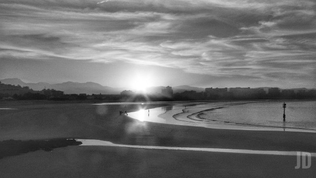 La imagen en blanco y negro muestra una playa durante el atardecer, con el sol asomando por detrás de edificios y montañas al fondo. La marea está baja, creando charcos y reflejos en la arena húmeda. Algunas personas caminan por la orilla, y el cielo está parcialmente cubierto por nubes finas que difuminan la luz.