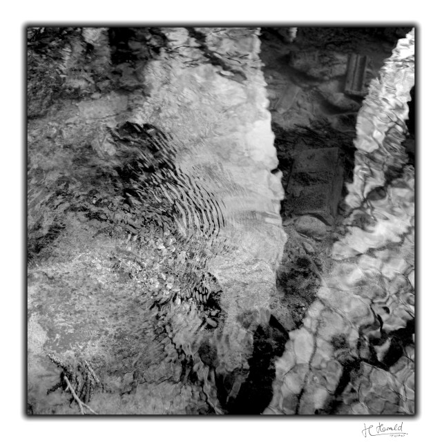 Square black-and-white image with a white frame. It shows the surface of a small stream with light and dark patches. In the centre, the water’s surface ripples slightly, and from the top right, the shadow of a tree falls across it. In this area, you can see the bottom of the stream, where there is, among other things, a brick and fragments of roof tiles. 