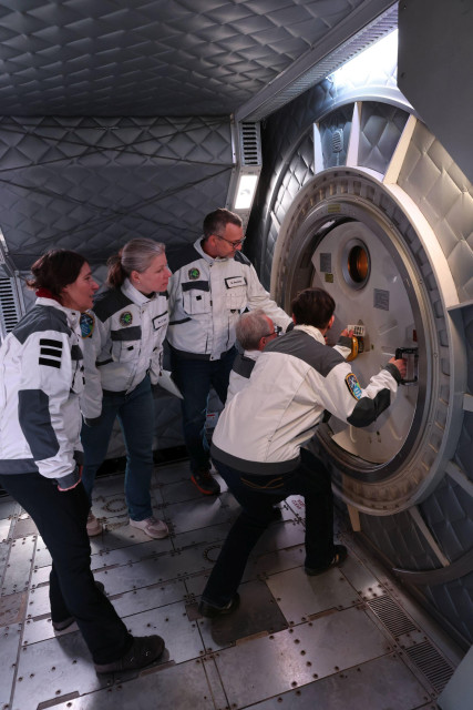 Fünf Personen (zwei Damen, drei Herren) vor einem spacigen Schott, wie in einer Raumstation. Sie scheinen zu versuchen, es zu öffnen.