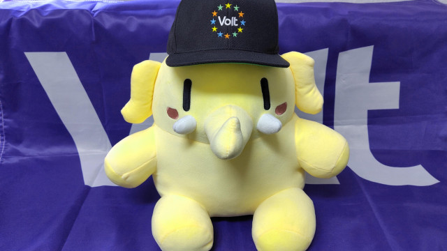 A yellow Mastodon mascot plushie, known as "Voltodon," sits centered against a vibrant purple Volt Europa flag. The plushie is round and soft, with small black vertical oval eyes, white tusks, and a short trunk. It wears a black baseball cap featuring the Volt logo: the word "Volt" in white text, surrounded by a circular crown of twelve stars in various colors (yellow, green, blue, and red).

The background is a deep purple fabric flag with the large, white, sans-serif letters "Volt" partially visible behind the plushie. The lighting is bright and even, highlighting the contrast between the sunny yellow of the Voltodon and the signature purple of the Volt brand. The overall vibe is friendly, cute, and politically engaged, symbolizing the intersection of the Volt Europa movement and the Fediverse community.