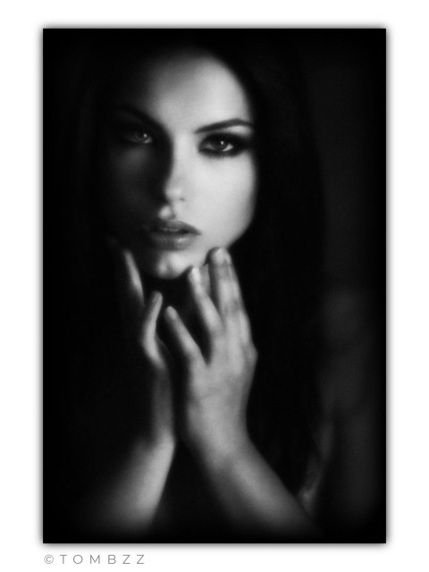 A moody, black-and-white analog photograph featuring a portrait of a young woman emerging from deep shadows. The model gazes directly into the lens with a piercing and mesmerizing look. Her dark, long hair blends into the black background. Her hands are raised to her face, with fingers resting gently against her chin and cheeks, creating a symmetrical frame for her lips. The lighting is soft, focused primarily on the center of her face, giving the image an ethereal, almost painterly quality. The shot is characterized by low contrast and a delicate blur typical of retro techniques.