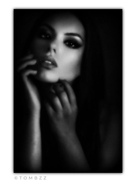 An artistic female portrait captured through traditional black-and-white photography. The woman's head is slightly tilted back and turned to the side, giving the pose a more dynamic yet dreamy expression. Her eyes are directed slightly upward and away from the camera, avoiding direct contact with the viewer. One hand rests near her lips while the fingers of the other elegantly trace her jawline, accentuating the neck. The interplay of light and shadow (chiaroscuro) is very distinct here—the right side of her face remains in darkness while the left is subtly illuminated. The entire image has a grainy texture and a very sensual, mysterious atmosphere.