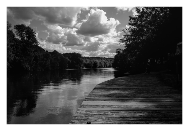 Daylight monochrome photography.
Contrasts over the river, crowded cloudy sky, dark banks, bright reflections on the river, a wooden dock at the foreground.