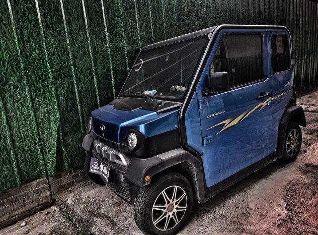 Bright blue smaller car against some metal panelling decorated with a photographic print of some grass.