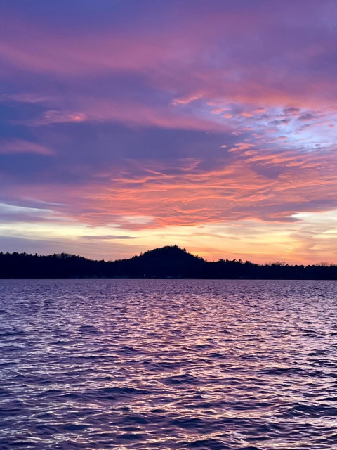 A sunset over a choppy lake. The sunset is orange at the horizon and lights the bottom of a large undulating cloud with bright red. Above this the clouds are a purplish color. The choppy water does not provide a symmetrical view, but instead resolves to a diffuse purplish color. Some nights the water is purple and this was one of those special nights. 