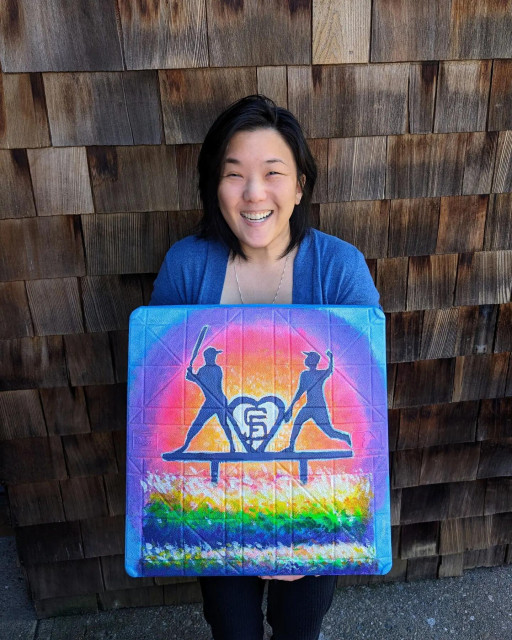 Joy holding up her painted baseball base for the San Francisco Giant's Pride Base Art campaign. It is a painting of a rainbow cake with two baseball players as cake toppers. One player is batting, one is pitching, and between is a heart with the SF Giants logo in it.