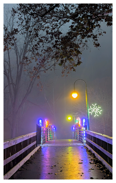 A wooden bridge on a misty night is illuminated by colorful holiday lights and an overhead streetlamp. Bare tree branches hang above, and the wet surface of the bridge reflects the glowing lights and festive decorations in the foggy background.