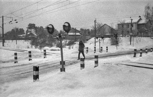 A grainy black-and-white photograph shows a quiet winter railway crossing covered in fresh snow. A lone person in a hooded jacket is walking along the road as it passes over the tracks, slightly hunched against the cold. Dark striped bollards line the crossing, creating a rhythmic pattern that contrasts with the white surroundings. Railway tracks cut across the scene, while overhead power lines stretch diagonally through the sky.

In the background, modest houses and bare trees are softened by the snowfall.
