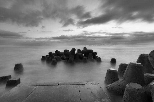 Black and white long-exposure seascape at Kadena Seawall, Okinawa, Japan. Concrete wave breakers emerge from smooth, misty water beneath dramatic, layered clouds.