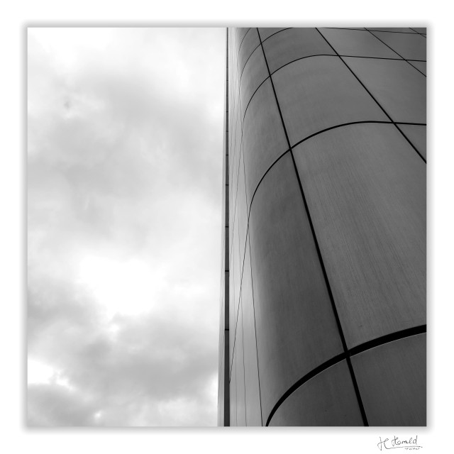 A square black-and-white photograph with a white border of the Silver Tower in Frankfurt am Main. Grey clouds are visible in the left-hand half (or, to be precise, about 49%) of the image. On the right, the view angles upwards along a rounded corner of the building. A dark line from a large window recess runs vertically through the centre of the image. No windows are visible, only the metal panels of the façade. 
