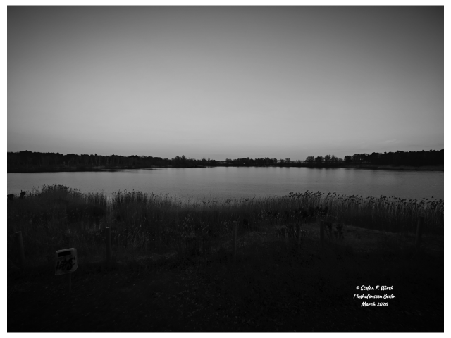 Artistic photography of lake Flughafenseen in Berlin, seen from its beach through a wide angle lens, 22 March 2026, © Stefan F. Wirth

Freshwater lakes interact with global warming in a complex and sometimes contradictory way, binding CO2 in their sediments but also releasing CO2 and methane through decomposition processes. They also warm up faster than their surroundings. On the other hand, the ecological conditions of the surrounding area are important for how "climate-friendly" a lake is. Berlin's Flughafensee (Airport Lake) lies in the middle of a forest (Jungfernheide Forest) and is partially protected by fences as a bird sanctuary. Therefore, an almost untouched shoreline vegetation thrives there, including marshy areas and reedbeds.