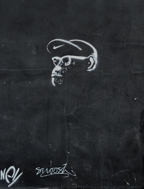 A photo of a white stencil graffiti on a dark wall depicting a gorilla head in profile with a Nike-branded cap. Small white graffiti tags can be seen at the bottom.