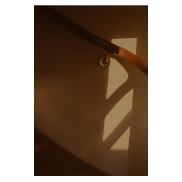 Ein Treppenaufgang. Quer über einen hölzernen Handlauf hat die Morgensonne einen rechtwinkligen Sonnenspot an die weiße Wand geworfen. 