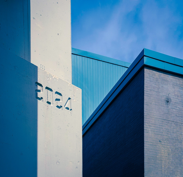 Small portion of a concrete bridge and a brick and aluminum siding building beside it. It’s all in hues of blues - even the sky behind. The bridge is marked with the year it was built (rebuilt in this case) which is 2024. The photo is all peaks and lines and angles. 